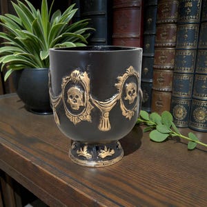 May include: Black decorative cup with gold accents, featuring skull designs within ornate frames. The cup has a pedestal base and is likely made of ceramic or a similar material. The cup is displayed on a wooden surface.