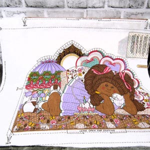 May include: A fabric panel with a pattern of a brown-skinned girl with brown hair wearing a purple dress and holding a white bird. The girl is lying on a bed with a floral pattern. The panel is surrounded by a white border with instructions for sewing. The text on the panel reads "Leave open for stuffing."