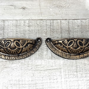 May include: Two antique-style drawer pulls with a bronze finish. Each pull is a half-moon shape with an ornate, symmetrical design. The pulls have a dark, aged appearance, with two holes for mounting.