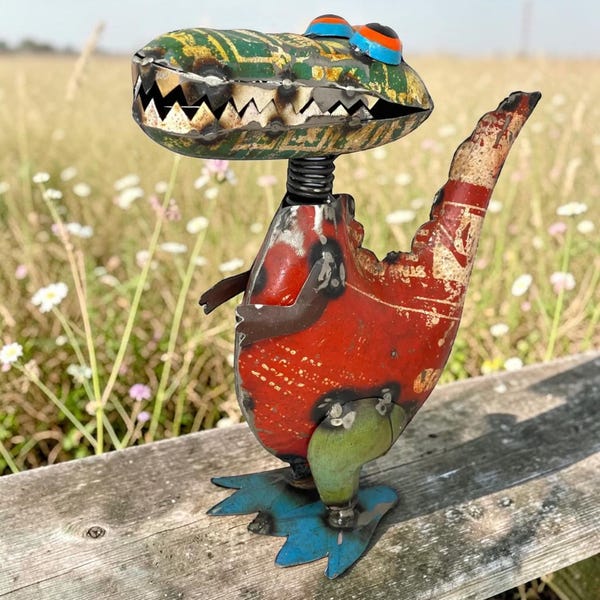 May include: A metal sculpture of a dinosaur crafted from recycled scrap metal. The head is green with yellow patterns, the body is red, the legs are green, and the feet are blue. It features a spring neck and large, colorful eyes.