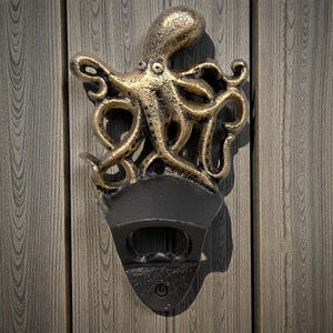 May include: A cast iron bottle opener featuring an octopus design. The octopus is finished in a gold color, while the bottle opener is black. The opener is mounted on a wooden surface.