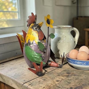 May include: A whimsical metal rooster sculpture with a red comb, yellow beak, and purple body. It holds a yellow sunflower and has green accents. The sculpture is on a wooden surface, with a ceramic pitcher and a bowl of eggs in the background.