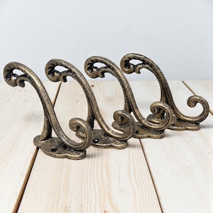 May include: Four antique-style bronze wall hooks. Each hook features a curved design with decorative swirls and a flat base for mounting. The hooks are arranged in a row on a light wood surface.