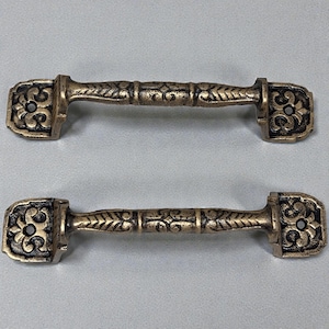 May include: Two antique-style bronze cabinet pulls. Each pull features a rectangular end with a floral design and a central handle with leaf-like detailing. The pulls are displayed against a neutral background.