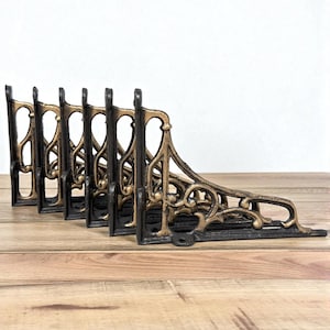 May include: Set of six black and gold shelf brackets. The brackets have an ornate, vintage design with curved lines and decorative flourishes. They are made of metal and are suitable for supporting shelves. The brackets are displayed on a wooden surface.