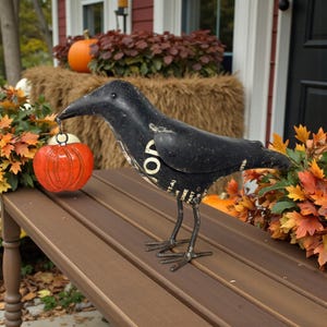 May include: A black metal crow figurine holding a small orange pumpkin. The crow has a distressed finish and is perched on a brown bench. Autumn leaves and pumpkins decorate the background, creating a fall theme.