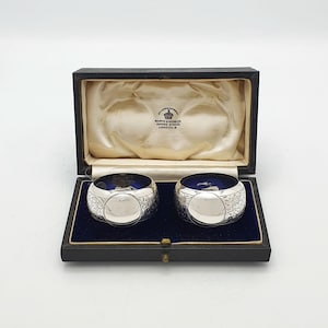 May include: A pair of silver napkin rings with a blue interior, presented in an open black box with a blue velvet lining. The napkin rings feature an engraved pattern. The box has a cream satin lining.
