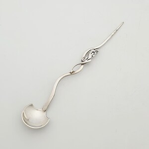May include: A silver ladle with an ornate handle and a small, rounded bowl. The handle features a decorative design with a curved shape and a pointed end. The ladle is likely used for serving sauces or other liquids.