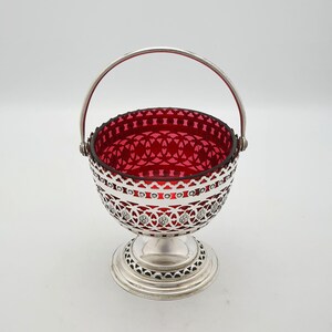 Op de afbeelding: Een vintage, verzilverde mand met een robijnrode glazen binnenkant. De mand heeft een sierlijk opengewerkt ontwerp rond de rand en de basis, met een bijpassende handgreep. De mand staat op een voet.