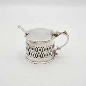 以下が含まれることがあります： 蓋とハンドルが付いた銀色の砂糖入れ。ボウルは側面に格子模様があり、緑色の内側が見えます。小さなスプーンが蓋の上にあります。ハンドルは装飾的で湾曲したデザインです。