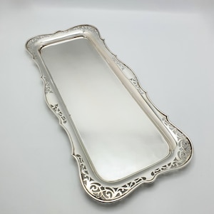 Könnte beinhalten: Ein rechteckiges silbernes Serviertablett mit verzierten, gerollten Kanten. Das Tablett hat eine reflektierende Oberfläche und dekorative Ausschnitte entlang des Randes. Das Tablett ist wahrscheinlich aus versilbertem Metall.
