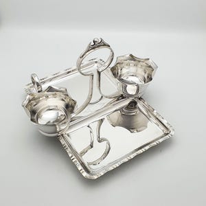 Może przedstawiać: Srebrny zestaw do serwowania z dwiema tacami i dwiema małymi miseczkami. Zestaw zawiera uchwyt ułatwiający przenoszenie. Miski mają falisty brzeg. Tace i miski są odblaskowe i wydają się być wykonane z metalu.