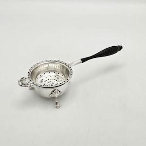 May include: A silver tea strainer with a polished finish, featuring a perforated bowl, three decorative shell-shaped feet, and a long, slender black handle. The rim of the bowl is detailed with a beaded pattern, and the handle is smooth.