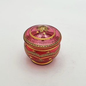 Puede incluir: Un pequeño tarro redondo con tapa, de color rosa vibrante. El tarro está adornado con intrincados detalles dorados, incluyendo bandas y motivos florales. La tapa presenta un diseño floral dorado. El tarro es probablemente de vidrio o un material similar.