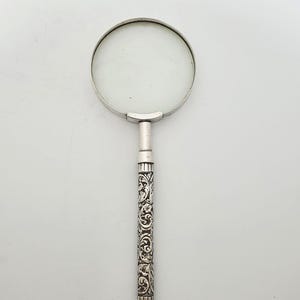 May include: A magnifying glass featuring a circular lens encased in a silver-toned metal frame. The handle is long and cylindrical, decorated with an intricate, embossed floral and scrollwork pattern in a dark, oxidised silver finish.