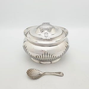 May include: A polished silver sugar bowl with a matching shell-shaped spoon. The bowl features a rounded body with vertical fluted detailing along the base and a lid with a central shell motif. A small circular hallmark is visible on the lid.