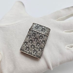 Könnte beinhalten: Ein rechteckiger Streichholzbehälter aus Silber mit einem komplizierten Filigranmuster aus feinen Drahtspiralen und kreisförmigen Motiven. Das Metallgehäuse verfügt über einen Klappdeckel und eine detaillierte metallische Oberfläche.