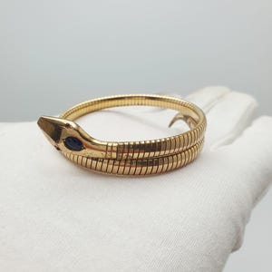 May include: A gold-toned snake bracelet featuring a flexible, segmented body that wraps around. The snake head is detailed with a dark blue oval-shaped stone for the eye and a small red stone accent near the snout, showcasing a polished finish.