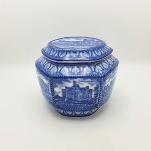 May include: A blue and white ceramic jar with a matching lid, featuring a hexagonal shape and detailed transferware illustrations of Warkworth Castle. The surface is decorated with intricate architectural scenes and repeating floral border patterns.