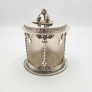 May include: A silver-coloured, cylindrical container with a lid. The lid is topped with a decorative floral design. The container has a detailed, ornate design around the rim and base, with a twisted central support.