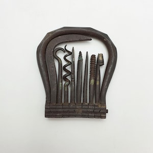 May include: A vintage folding multi-tool set featuring a dark, oxidised metal frame that encloses several distinct implements, including a corkscrew, a hook, and various pointed metal shafts, all arranged in a compact, fan-like configuration.