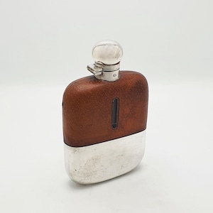 Peut inclure: Une flasque de poche composée d'une partie supérieure gainée de cuir marron avec une fenêtre de visualisation verticale et d'une partie inférieure en métal argenté poli. La flasque est surmontée d'un bouchon arrondi et d'une charnière.