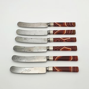 May include: A set of six butter knives with silver-toned metal blades and rectangular handles featuring a brown and white banded agate stone pattern. Each knife has a rounded tip and a polished metal bolster connecting the blade to the handle.