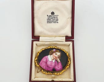 May include: An oval brooch featuring a hand-painted portrait of a person in a pink shirt against a black background, set in a gold-tone ornate frame. The brooch is housed in a maroon presentation box with text reading By appointment to H.M. THE QUEEN.
