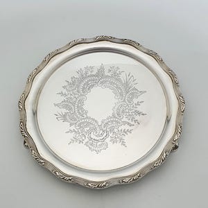 Op de afbeelding: Een rond zilverkleurig metalen dienblad met een geschulpte, decoratieve rand. Het midden van het dienblad is voorzien van een ingewikkeld, gegraveerd bloemen- en krulpatroon in een cirkelvormig ontwerp. Het oppervlak is reflecterend en gepolijst.