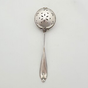 May include: A silver-toned metal tea infuser spoon featuring a spherical, hinged head with a perforated floral pattern and a long, slender handle with decorative embossed detailing at the base. The object has a polished, reflective metallic finish.