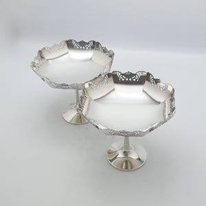 Może przedstawiać: Dwie srebrne misy na nóżkach z ozdobnymi, falistymi krawędziami. Misy mają płytki, okrągły kształt i osadzone są na krótkiej, zaokrąglonej nóżce. Krawędzie zdobione są dekoracyjnym ażurowym wzorem. Misy prezentowane są na gładkim, białym tle.