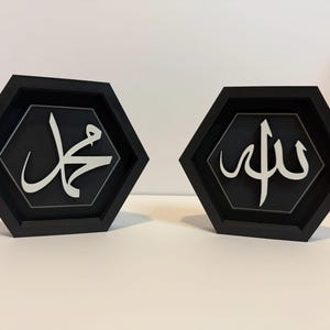 Op de afbeelding: Twee zwarte zeshoekige frames met witte Arabische kalligrafie. Het linker frame zegt "Muhammad" en het rechter frame zegt "Allah".