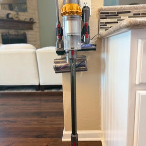 K&ouml;nnte beinhalten: Ein lila und gelber Dyson-Akkusauger ist an einer Wandhalterung mit Ladestation befestigt. Der Staubsauger befindet sich in vertikaler Position mit der Reinigungsd&uuml;se unten. Der Staubsauger ist mit "Dyson V15 Detect" beschriftet.