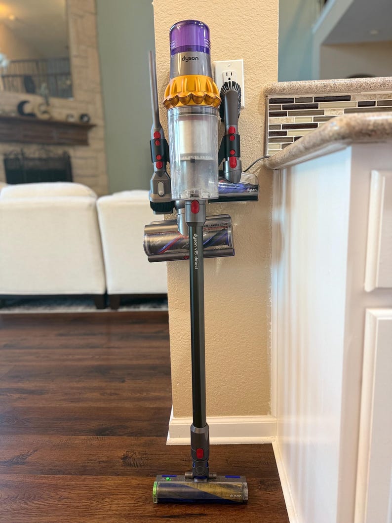 K&ouml;nnte beinhalten: Ein lila und gelber Dyson-Akkusauger ist an einer Wandhalterung mit Ladestation befestigt. Der Staubsauger befindet sich in vertikaler Position mit der Reinigungsd&uuml;se unten. Der Staubsauger ist mit "Dyson V15 Detect" beschriftet.