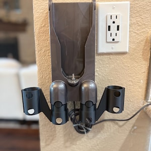 K&ouml;nnte beinhalten: Eine graue Wandhalterung mit zwei schwarzen Haltern f&uuml;r einen Staubsauger. Die Wandhalterung hat einen Schlitz f&uuml;r das Ladekabel des Staubsaugers. Die Wandhalterung ist an einer Wand mit einer wei&szlig;en Steckdose befestigt.
