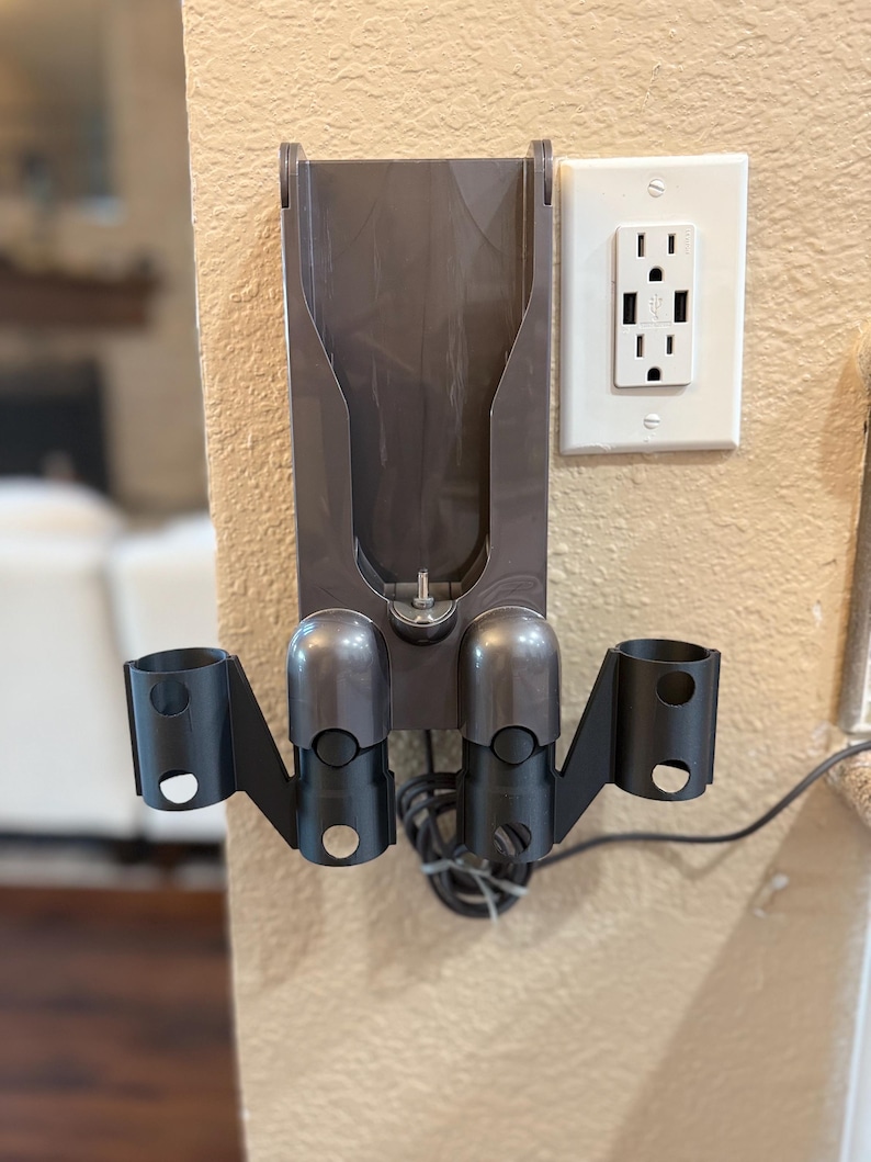 K&ouml;nnte beinhalten: Eine graue Wandhalterung mit zwei schwarzen Haltern f&uuml;r einen Staubsauger. Die Wandhalterung hat einen Schlitz f&uuml;r das Ladekabel des Staubsaugers. Die Wandhalterung ist an einer Wand mit einer wei&szlig;en Steckdose befestigt.
