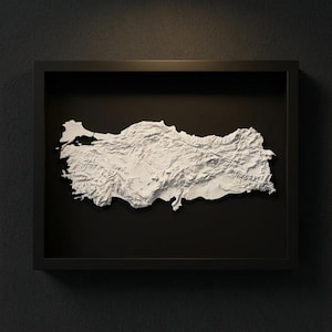 May include: A framed, three-dimensional relief map of Turkey, rendered in white against a black background. The map is detailed, showing the country's topography within a black, rectangular frame. The artwork is mounted on a dark gray wall.