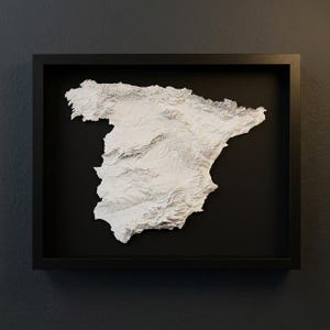 May include: A framed, three-dimensional relief map of Spain, rendered in white against a black background. The map is detailed, showing the country's topography. The frame is black, and the artwork is mounted on a dark gray wall.