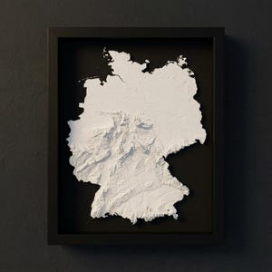 Könnte beinhalten: Eine gerahmte, dreidimensionale Reliefkarte von Deutschland. Die Karte ist weiß, mit detaillierten topografischen Merkmalen, vor einem schwarzen Hintergrund in einem schwarzen Rahmen. Das Kunstwerk ist eine moderne, minimalistische Darstellung der Landschaft des Landes.