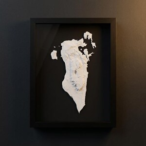 May include: A framed, three-dimensional relief map of Bahrain. The map is white and set against a black background within a black frame. The relief shows the island's topography, with varying heights and textures.