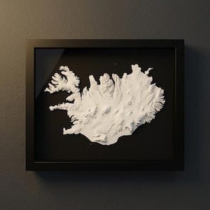 May include: A framed, three-dimensional white relief map of Iceland, set against a black background. The map is detailed, showing the island's topography. The frame is black and rectangular, mounted on a dark gray wall.