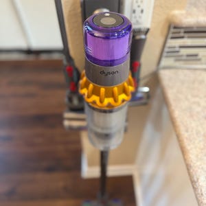 K&ouml;nnte beinhalten: Ein lila-goldener Akku-Staubsauger mit dem Schriftzug "dyson" an der Seite, der an einem Wandladeger&auml;t befestigt ist.
