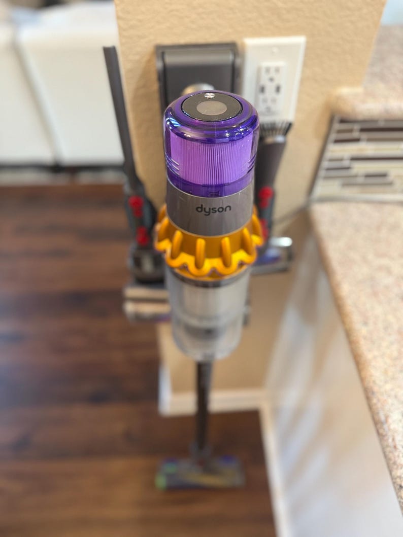 K&ouml;nnte beinhalten: Ein lila-goldener Akku-Staubsauger mit dem Schriftzug "dyson" an der Seite, der an einem Wandladeger&auml;t befestigt ist.