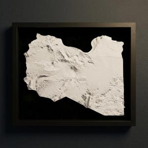May include: A framed, three-dimensional relief map of Libya, rendered in white against a black background. The map is encased in a dark wooden frame, showcasing the country's geographical features with textured detail. The artwork is mounted on a dark gray wall.