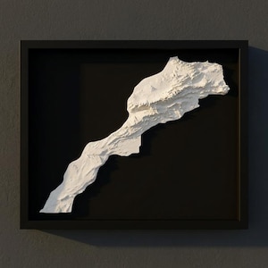 Puede incluir: Un mapa en relieve blanco enmarcado de Marruecos sobre un fondo negro. El mapa es tridimensional, mostrando la topografía del país. El marco es negro y la obra está montada en una pared gris oscura.
