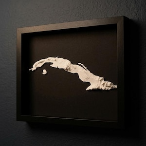 May include: A framed artwork with a white relief map of Cuba set against a black backdrop. The map is enclosed in a square, black wooden frame, providing a strong contrast. The artwork is mounted on a dark blue wall.