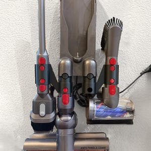 K&ouml;nnte beinhalten: Eine graue Wandhalterung mit einem Dyson-Staubsauger und Zubeh&ouml;r. Die Wandhalterung hat drei Steckpl&auml;tze f&uuml;r Zubeh&ouml;r und einen Steckplatz f&uuml;r den Staubsauger. Das Zubeh&ouml;r umfasst einen Fugend&uuml;se, eine Staubb&uuml;rste und einen Mini-Motor-Kopf. Der Mini-Motor-Kopf hat einen roten und blauen Anti-Verwicklungs-Kamm.