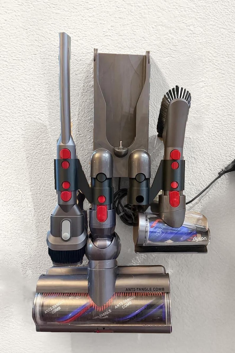 K&ouml;nnte beinhalten: Eine graue Wandhalterung mit einem Dyson-Staubsauger und Zubeh&ouml;r. Die Wandhalterung hat drei Steckpl&auml;tze f&uuml;r Zubeh&ouml;r und einen Steckplatz f&uuml;r den Staubsauger. Das Zubeh&ouml;r umfasst einen Fugend&uuml;se, eine Staubb&uuml;rste und einen Mini-Motor-Kopf. Der Mini-Motor-Kopf hat einen roten und blauen Anti-Verwicklungs-Kamm.