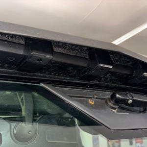 K&ouml;nnte beinhalten: Nahaufnahme eines schwarzen Scheibenwischerarms, der an der Windschutzscheibe eines Fahrzeugs befestigt ist. Der Wischerarm befindet sich in der oberen Position und ist mit einem schwarzen Wischerblatt verbunden. Der Wischerarm ist an einer schwarzen Metallhalterung befestigt, die an der Windschutzscheibe befestigt ist.