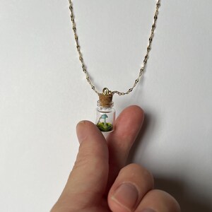 Puede incluir: Un collar de cadena dorada con un pequeño colgante de frasco de vidrio. El frasco contiene un hongo en miniatura y musgo verde.